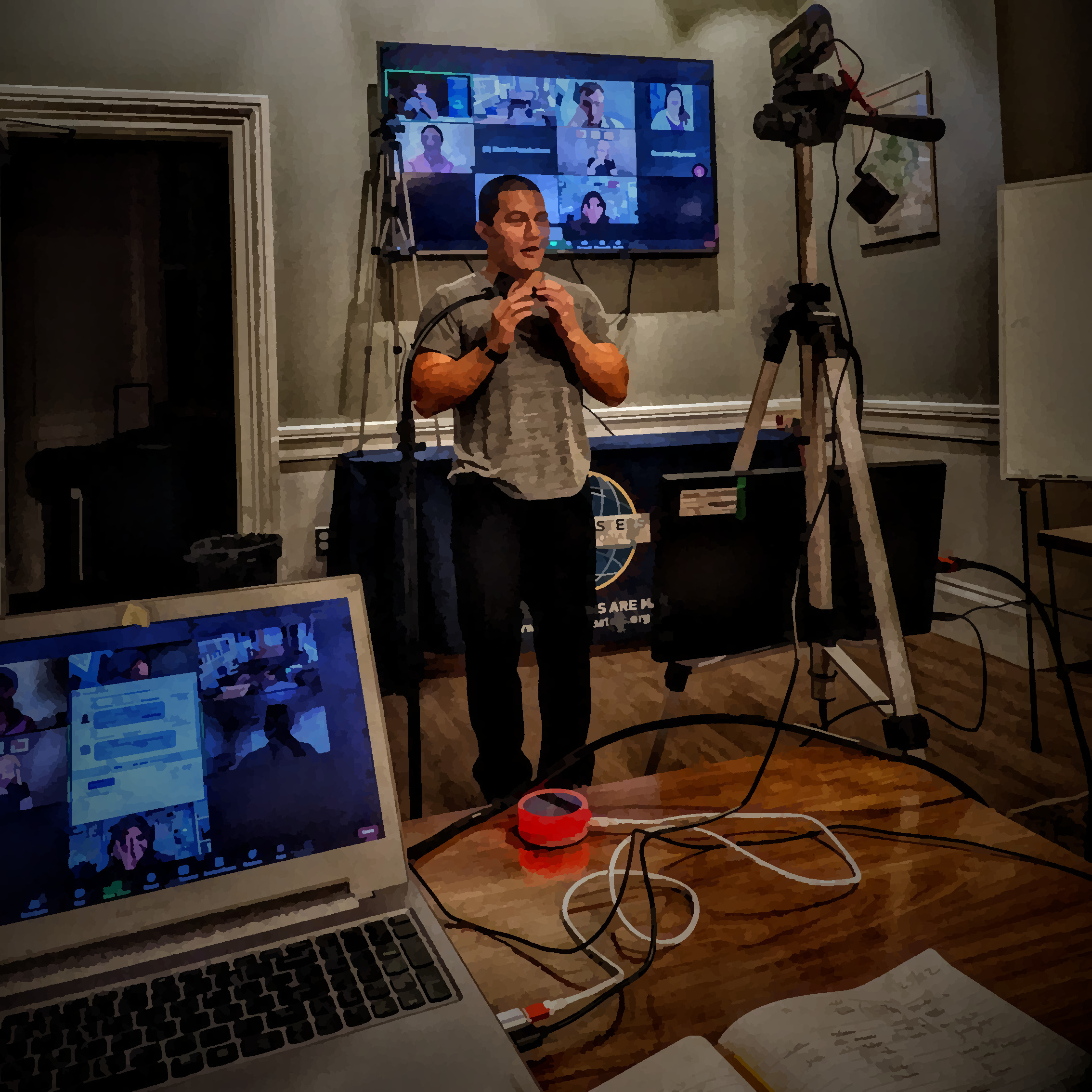 A man stands in front of a camera, with his hands gesturing in a triangle that points towards his face. In the background, we see a gallery of Zoom participants and in the foreground we see a camera and a laptop with the Zoom meeting running. A red "on-air" light glows on the table next to the laptop.